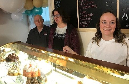 Three people standing behind a menu board in front of a glass case filled with cupcakes