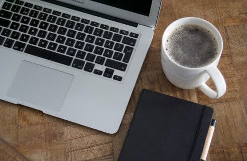Wood table with an open laptop computer, white mug of black coffee and a black notebook with pen