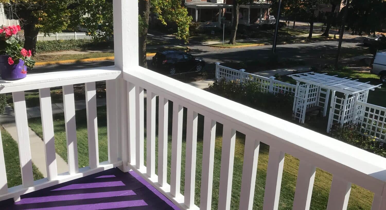 The corner of an upper outdoor balcony with white railings and purple flooring overlooking a yard
