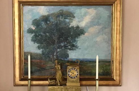 A painting of a landscape with a gold frame overtop a side table with candlesticks and gold antique clock piece