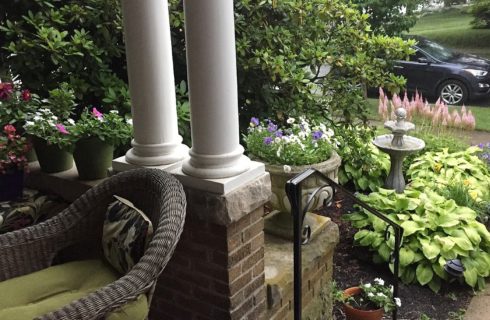 Corner of a front porch with brick wall, white columns, wicker chair and lush garden beds