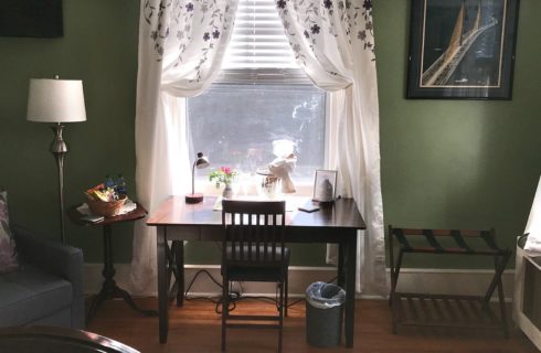 Brown writing desk in front of a large window with white floral curtains in a green guest room