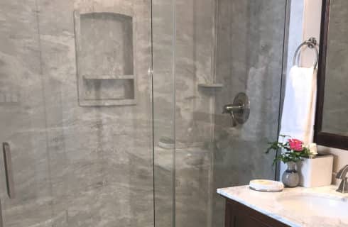 Large stand up shower with glass doors next to single vanity with marble countertop and large framed mirror