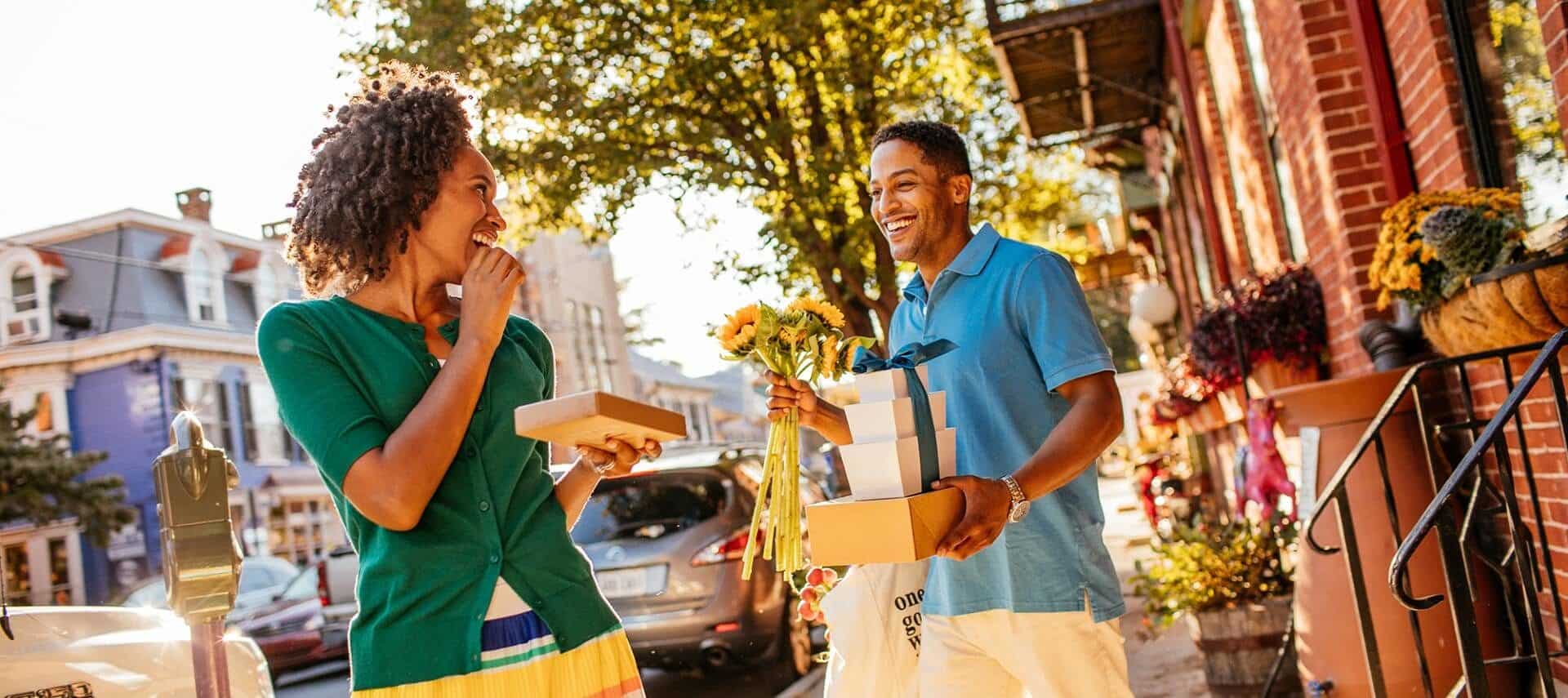 A man and woman smiling and looking at each other while holding packages standing outside on a downtown street