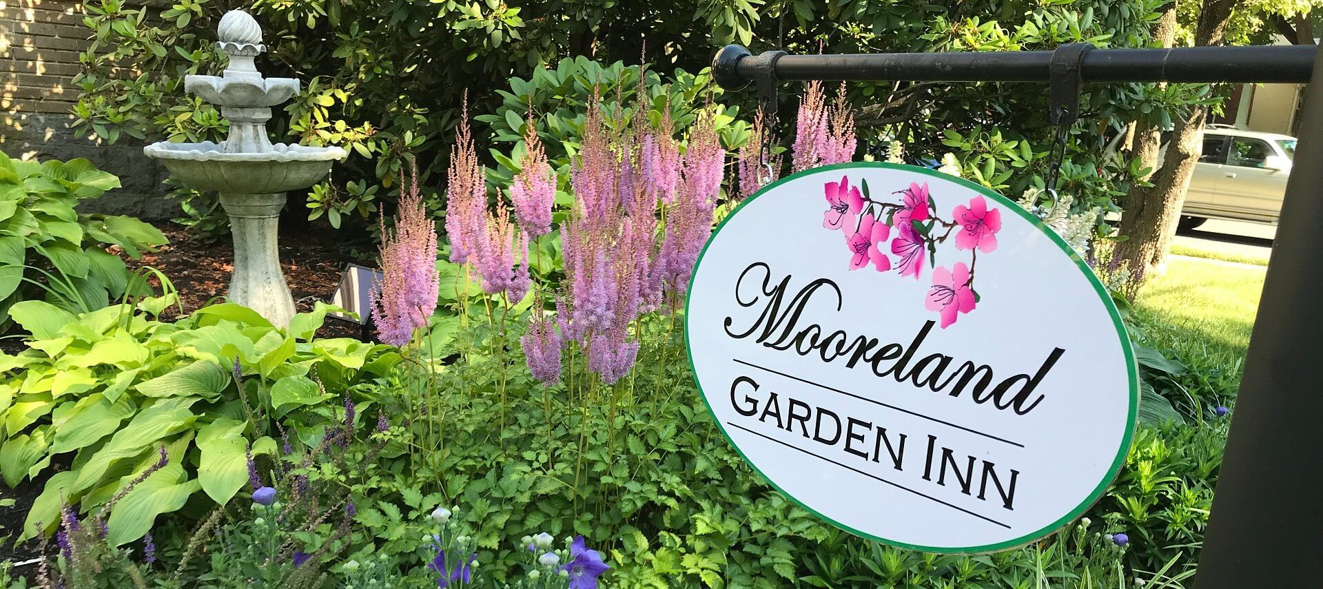 White oval business sign hanging on a black frame over a lush garden bed of flowers and water fountain