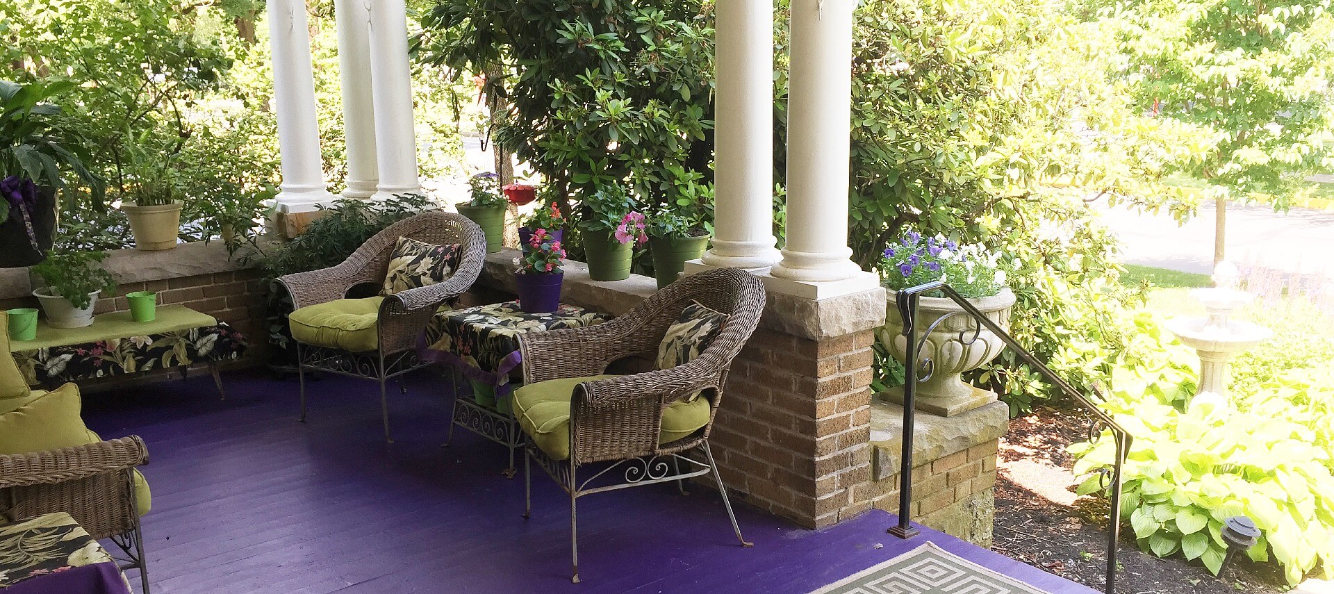 Spacious front porch of a home with wicker patio furniture, white columns and lush flowers and landscaping